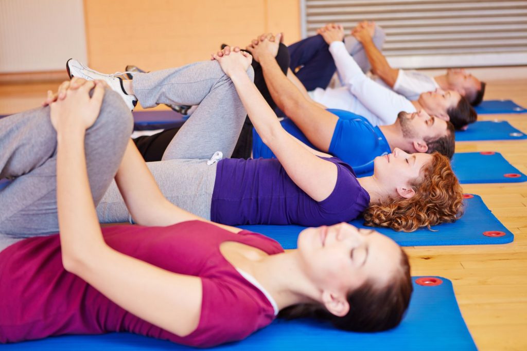 a group of 5 men and women laying on their backs holding one knee as part of a Feldenkrais exercise in a big room